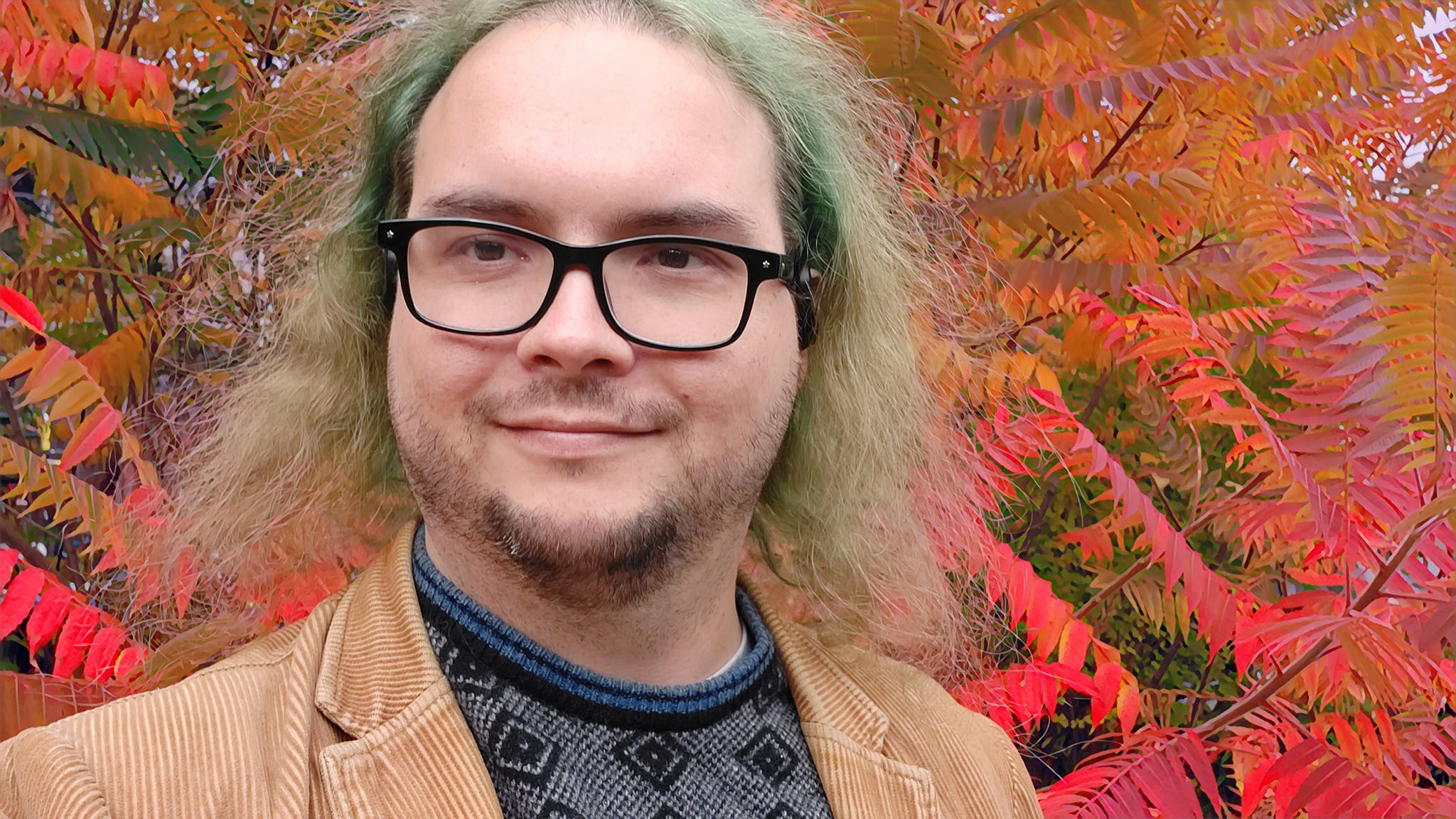 A man with long green hair stands in front of red and orange sumac leaves